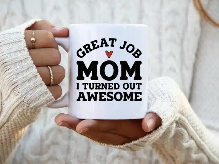 Close up of a woman's hands holding a white coffee mug with black font that says "Great job mom, I turned out awesome"