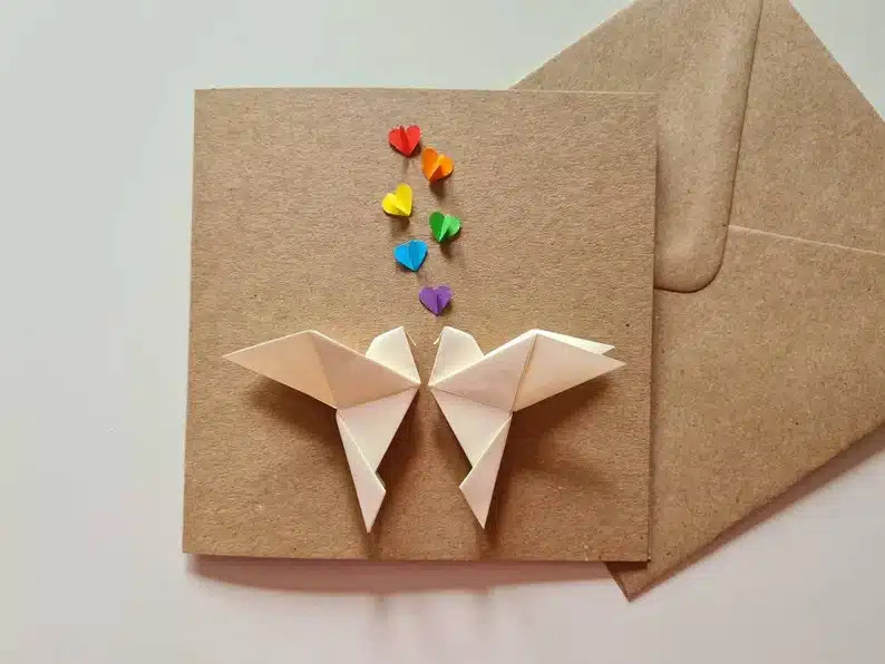 Brown card with two white origami lovebirds and rainbow hearts above. 