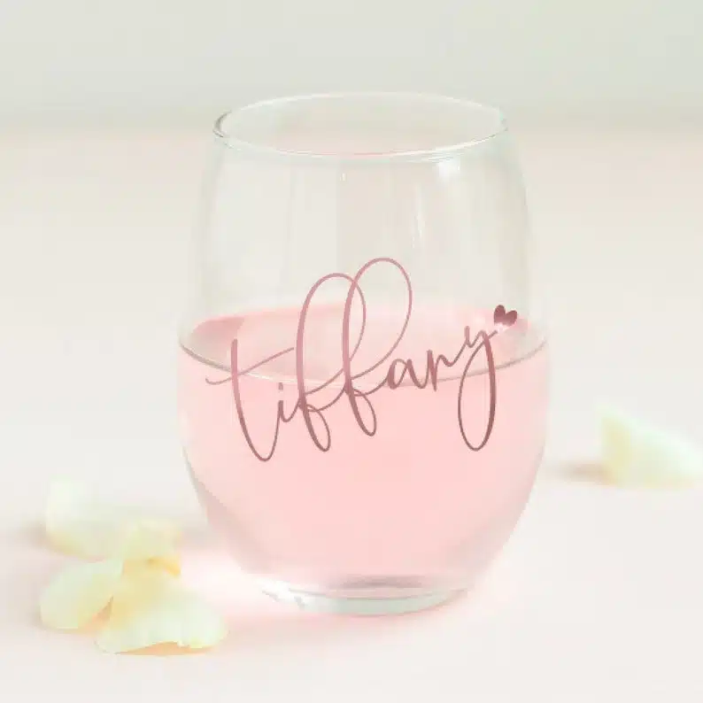 Clear stemless white glass with light pink font that says Tiffany on it with a light pink drink in the wine glass. 