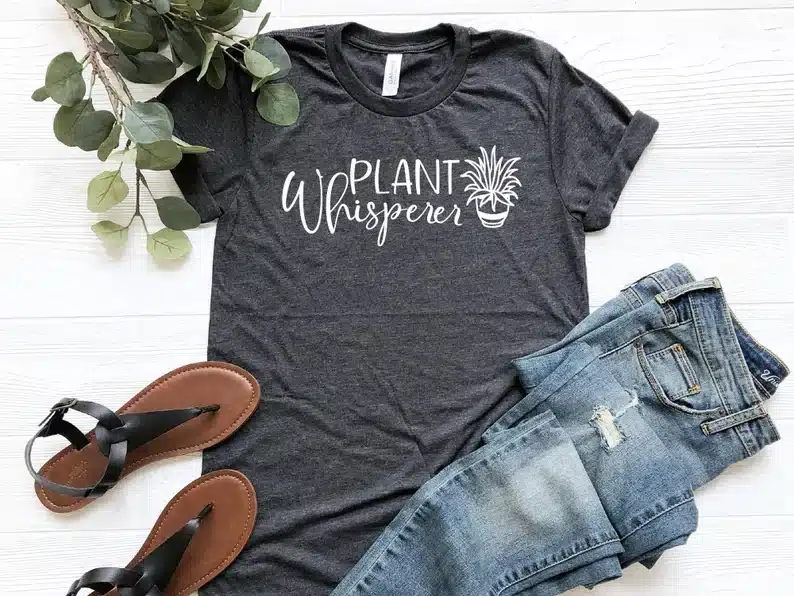 Grey t-shirt on a white background floor with white font that says plant whisperer with a plant, a pair of jeans beside it and brown sandals. 