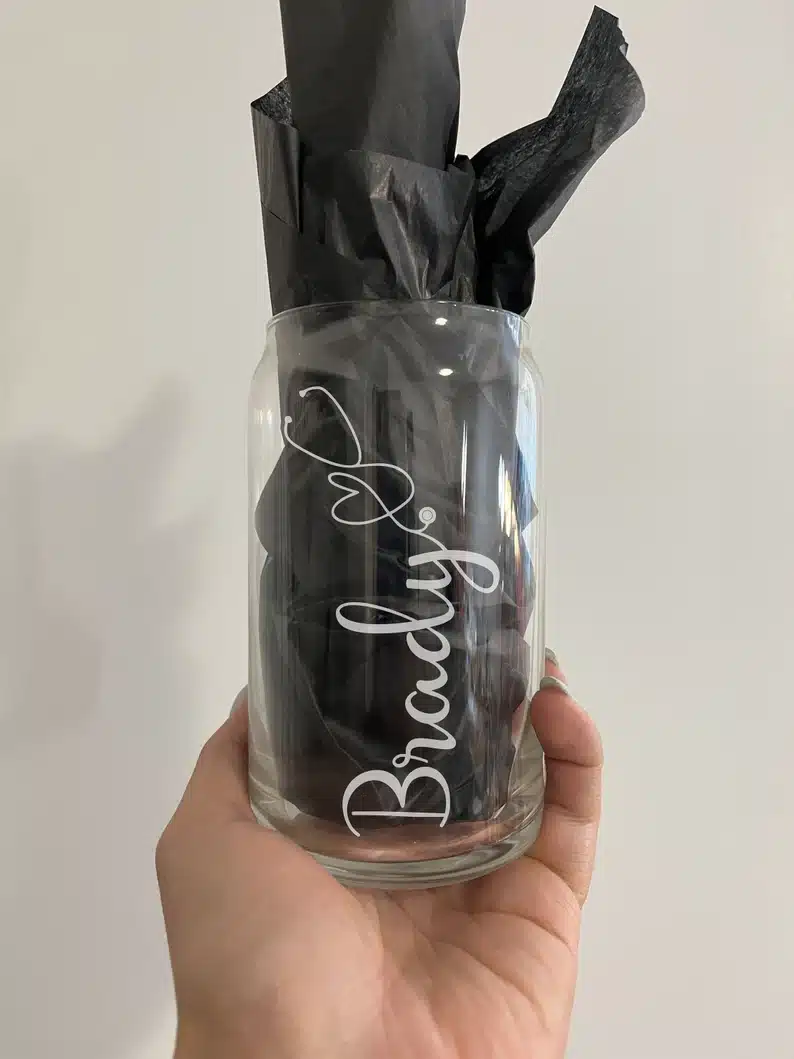 Hand holding a clear glass with black tissue paper in it with the name Brady etched into it. 