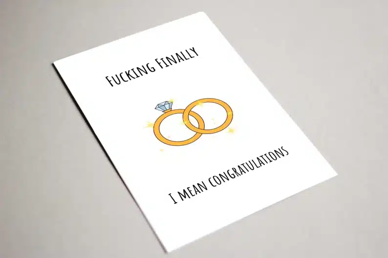 White card with black font that says "fucking finally I mean congratulations" with two gold rings on it. 
