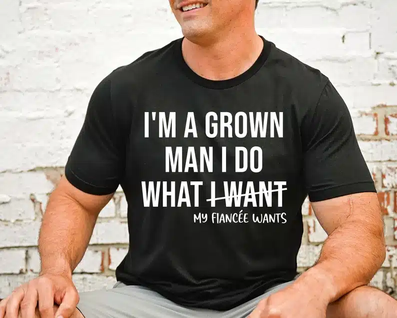 Man wearing a black t-shirt with white print that says "I'm a grown man I do what I want , I want being crossed out and My fiancee wants below. 