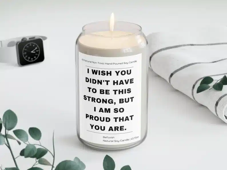 Clear can shaped clear glass with a white candle in it and a white label on it with black font that says "I wish you didn't have to be this strong, but I am so proud that you are" 