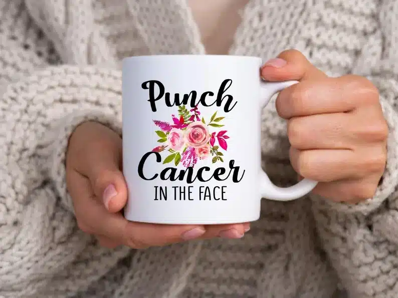 Close up of hands holding a white coffee mug that says "Punch cancer in the face" with pink flowers on it. 