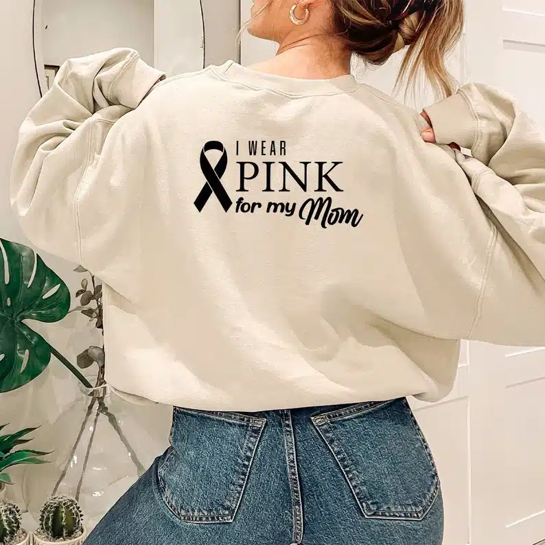 Back of a woman wearing a white long sleeve hoodie with black of it that says I wear pink for my mom with a ribbon. 