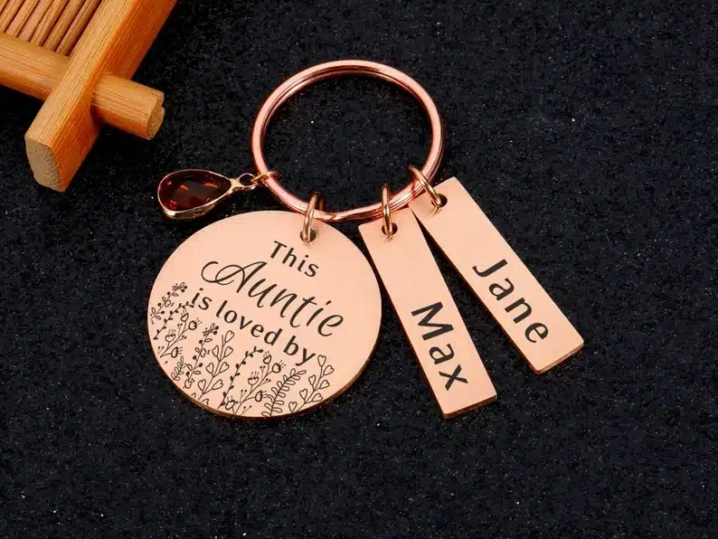 Rose gold keychain with a round charm that has black font that says This auntie is loved by and two rectangle charms each with black font that says Max and the other Jane. 