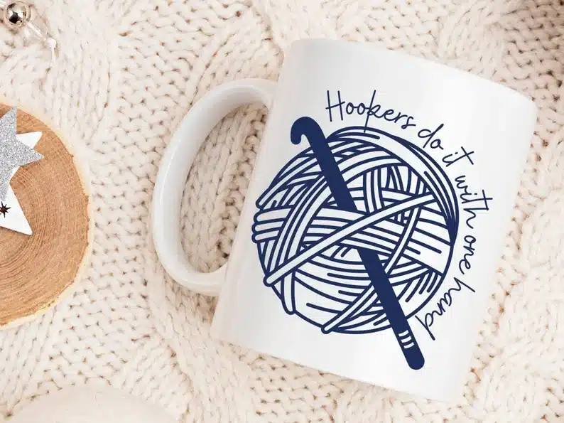 White coffee mug with nay font that says "hookers do it with one hand" with a yarn picture with a hooker needle. 