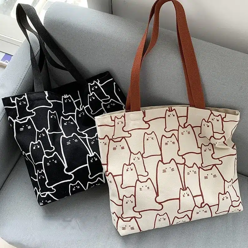Two large tote bags, one black with whtie outlines of cats all over it, the other cream with brown cats outlined all over it. 