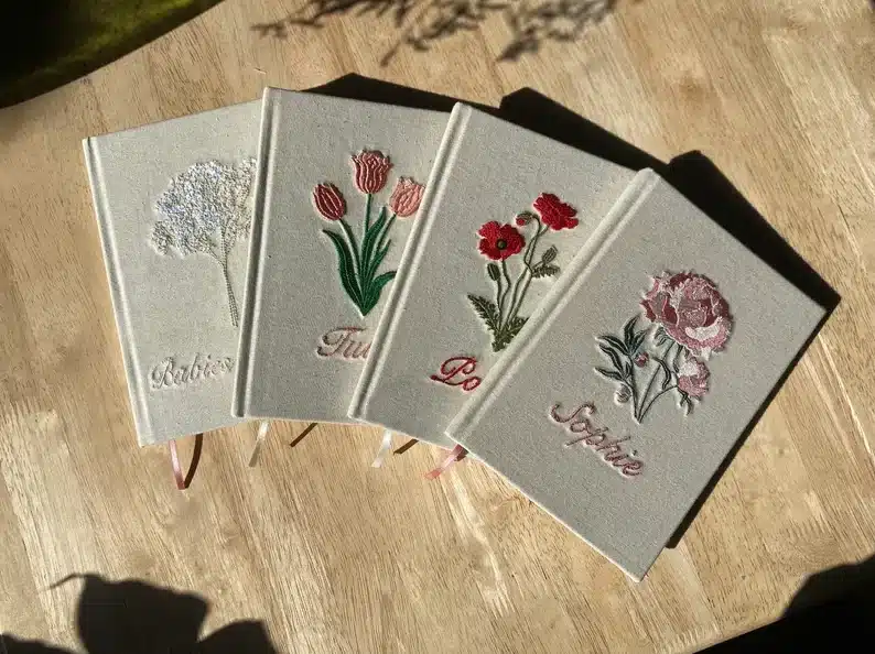 Four white notebooks each with a different flower on it and a name below. 