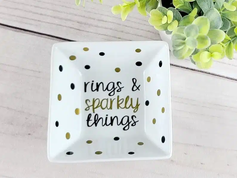 Above view of a square jewelry dish with gold and black spots all over it and black and gold font that says "rings & sparkly things:. 