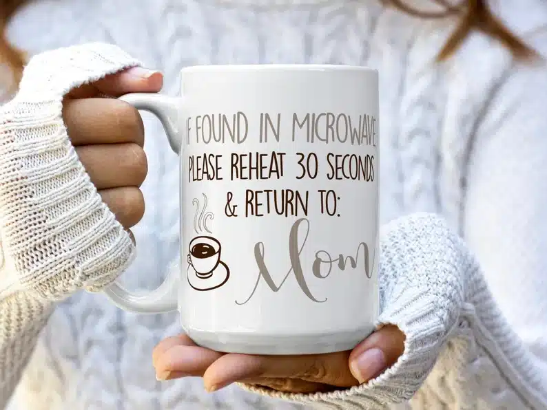 20 Mother’s Day Gift Ideas Under $20: close up of woman holding a white coffee mug that has various shades of brown font that says "If found in microwave please reheat 30 seconds & return to mom"