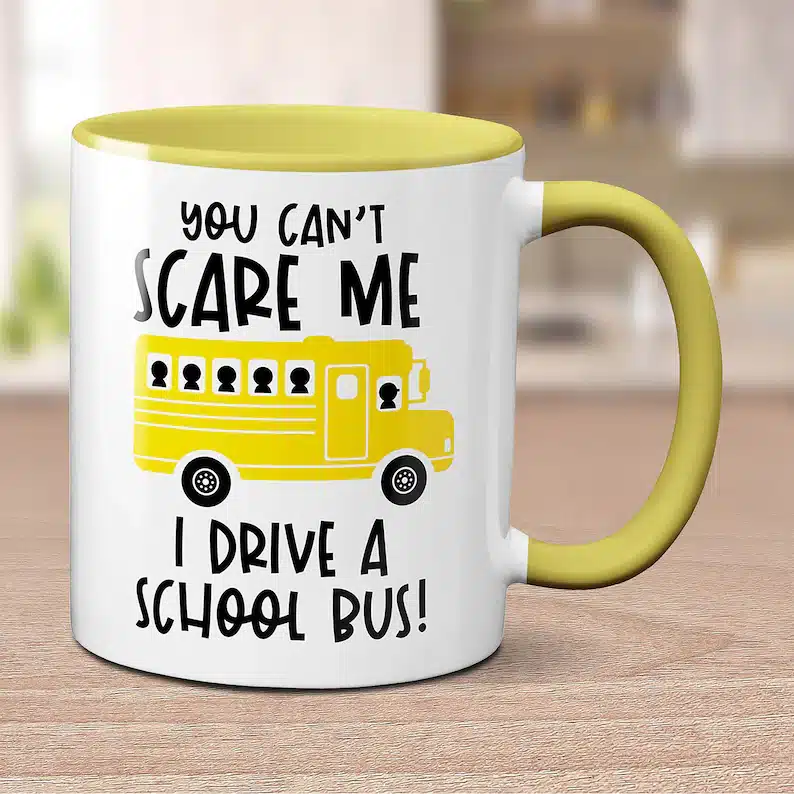 White coffee mug with a yellow handle with black font that says "You can't scare me, I drive a school bus!" with a yellow school bus on it. 