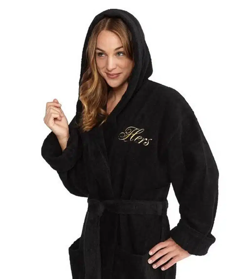 Relaxing Mother's Day Gifts: Woman wearing an all black hooded bath robe monogrammed with white font that says "hers". 