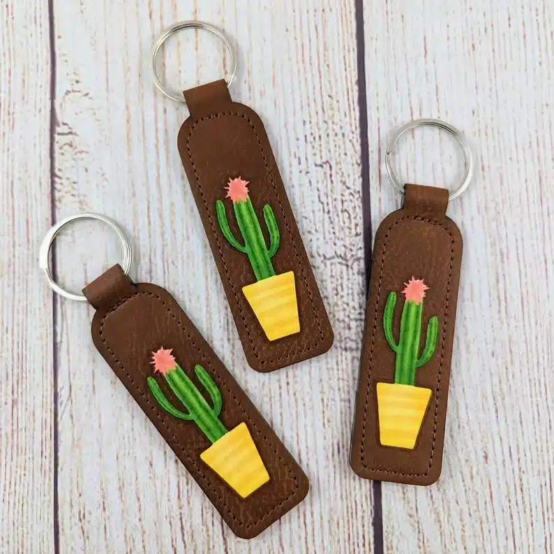Three leather brown cactus key fob, all with yellow pots and a green cactus with pink flower on top. 