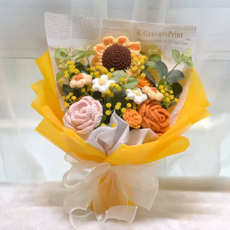 Crocheted floral bouquet with sunflower, roses, and small yellow flowers wrapped in white and yellow paper. 