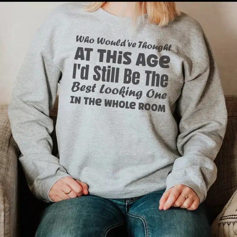 Neck down view of a woman wearing a light grey long sleeve sweatshirt with black font that says "Who would've though at this age I'd still e the best looking on in the whole room" 