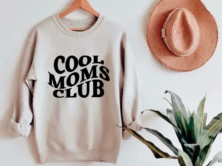Tan long sleeve sweatshirt with black font that says Cool moms club hanging beside a straw hat and a green plant. 