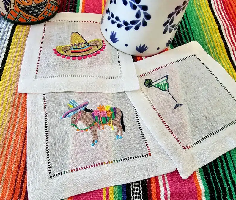 Three white embraided linen napkins, one with a sombrero, one with a green margarita, and one with a donkey wearing a sombrero. 