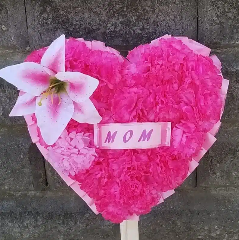 Heart shaped wreath with pink flowers all over it and one large pink lily with a rectangle sign that says MOM on it. 