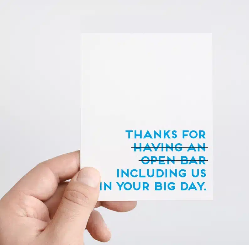 Close up of a hand holding a white wedding card with bright blue font that says Thanks for (having an open bar - crossed out) including us in your big day