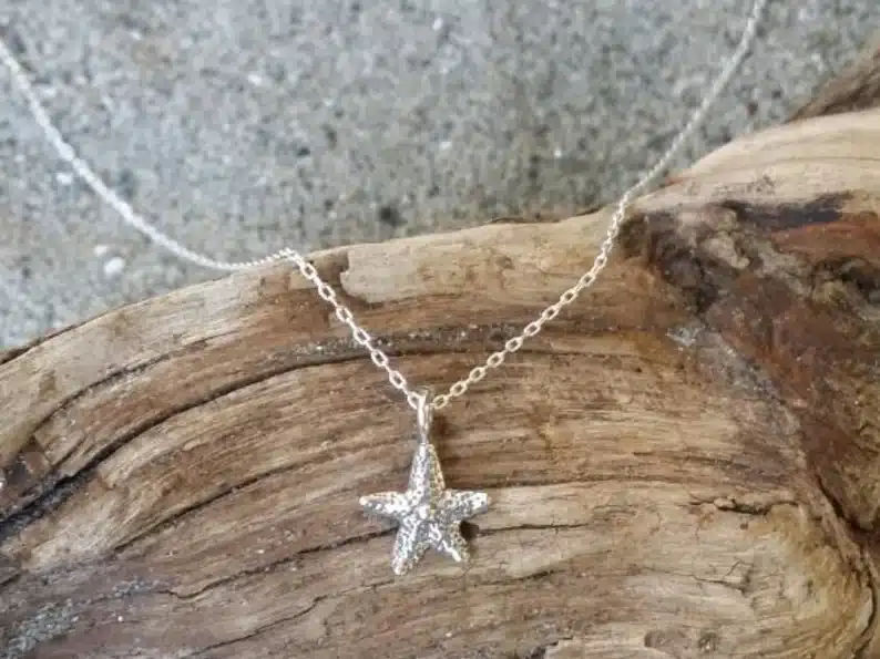 Silver necklace chain with a silver starfish charm laying on top of driftwood on a sandy beach. 