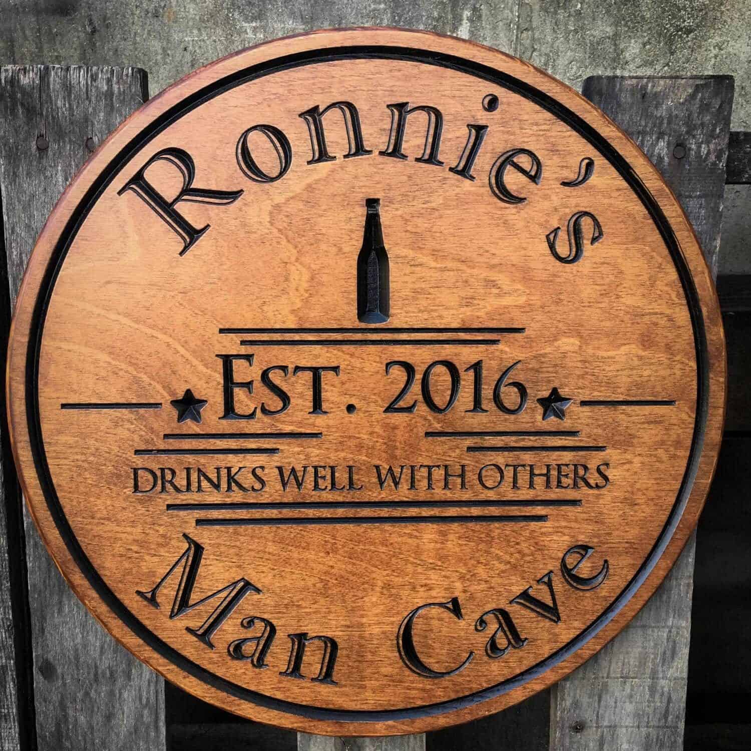 Round wooden sign with black print that says Ronnie's Est. 2016 Drinks well with others. Man Cave. 