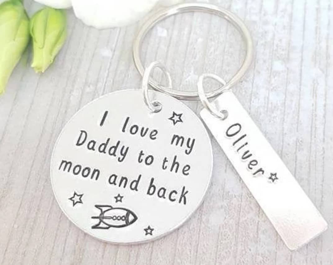 Silver keyring with a round charm that says "I love my daddy to the moon and back" with a rocket on it. Another rectangle long charm that says Oliver on it,