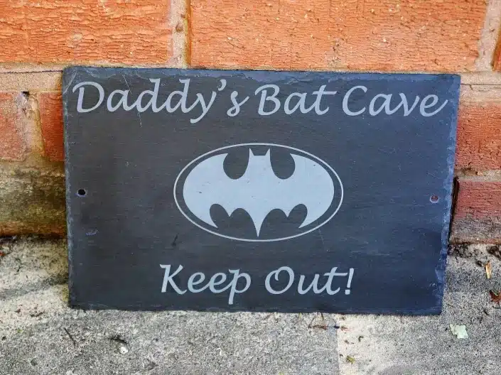 Black wooden sign that says Daddy's bat cave keep out! With the batman symbol on it in grey. 