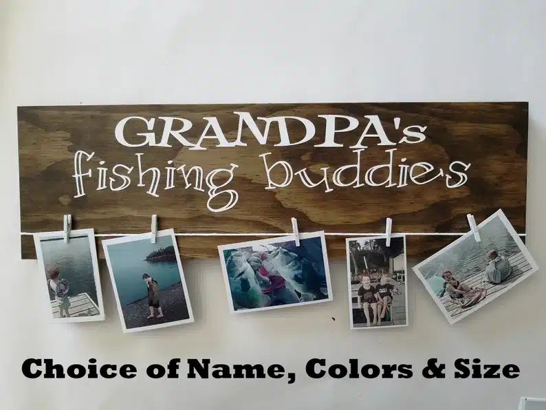 Wooden rectangle sign, with white font that says "Grandpa's fishing buddies" with white clothespins holding various photos of kids fishing. 