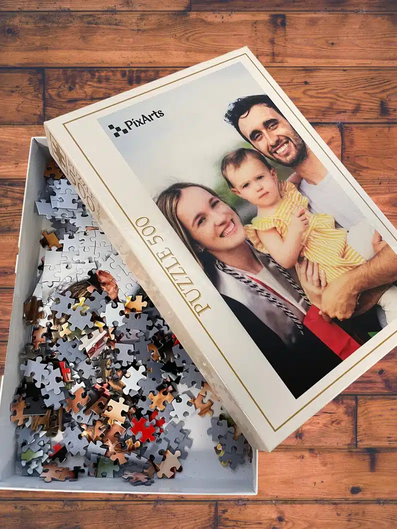 Open box reviling puzzle pieces, top of box has a family photo of a husband, wife and little girl. 