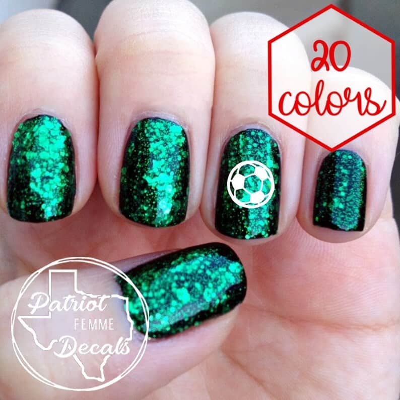 Close up of a woman's nails that are all glittered and green with a soccer ball sticker on one of them. 