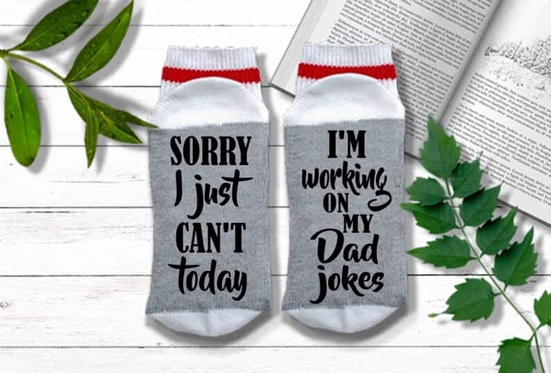 Funny Gift Ideas For Funny Father's Day - Light grey socks with white tips with black font that says "Sorry I just can't today" on one sock and "I'm working on my dad jokes" on the other.