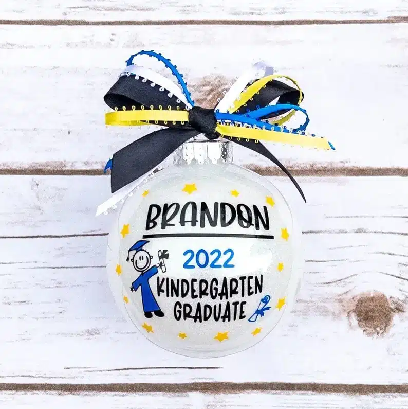 White round glass ornament with blue, white, yellow, and black ribbon on top with font on the ball that says Brandon 2022 Kindergarten Graduate. 