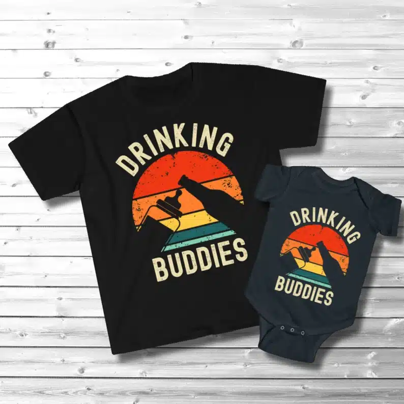 Matching black t-shirt and baby onesie. Both saying Drinking buddies on it with white font, a sunset with a silhouette or a milk bottle and beer bottle. 