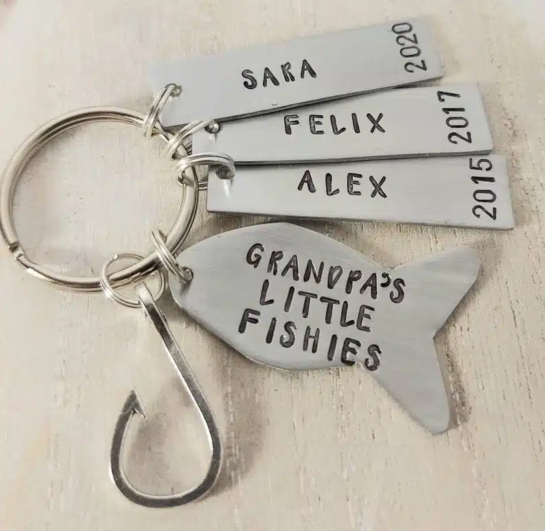 Silver keychain with a silver fishing hook on it and a fish shaped charm that says Grandpa's little fishies, with 3 other rectangle charms with names and birthdates on them. 