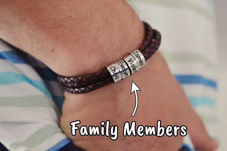 Close up of a mans wrist showing off a brown leather with four silver beads with names on them. 