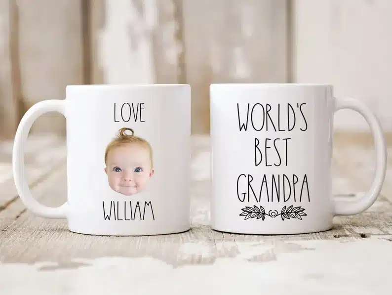 Two white coffee mugs shown, one that says Love William with a baby face on it and the other says World's best grandpa. 