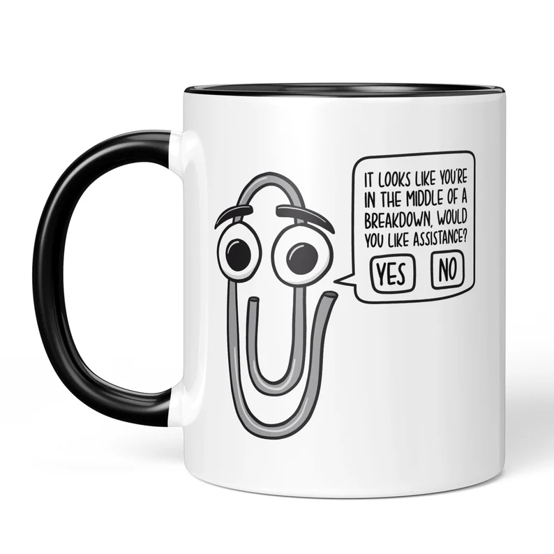 White coffee mug with black inside and black handle. Paperclip from word prefect on it that says it looks like you're in the middle of a breakdown who you like assistance. 