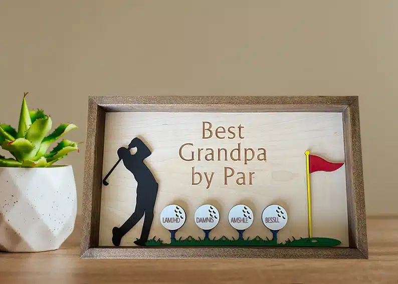 Wooden artwork with a man golfing and four white gold balls beside him each with a grandchild name on it. Above it says Best Grandpa by par. 