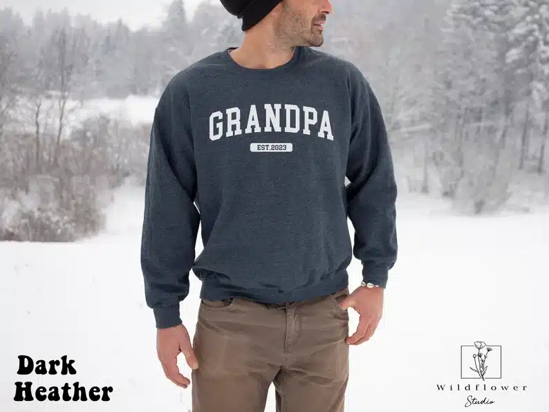 Fathers Day Gifts for Grandpa From Baby - Man standing outside in the snow wearing a dark grey long sleeve sweatshirt that says Grandpa est 2023 in white text. 