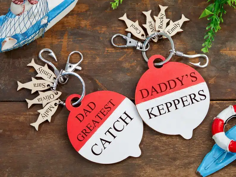 Two fishing bobbers one says  dad greatest catch and daddy's keepers. with little silver fish keychains with names on them. 