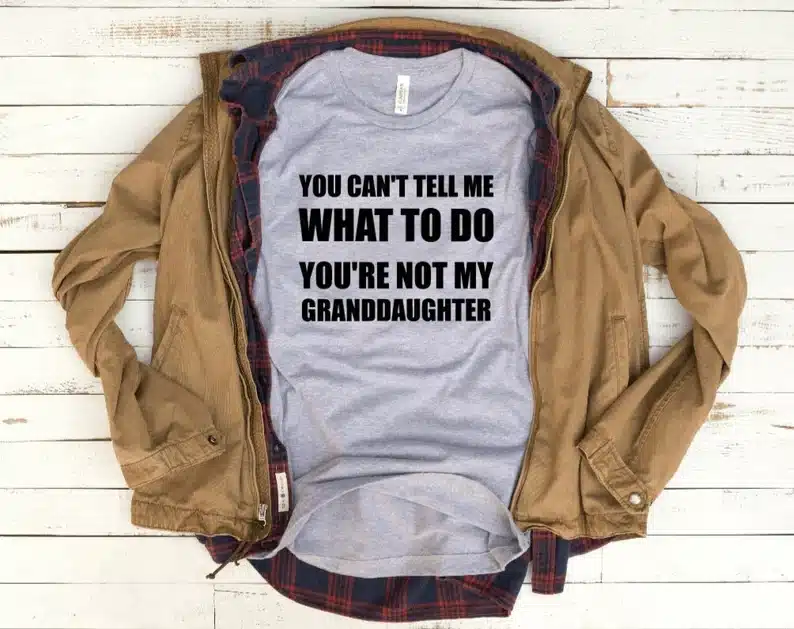 Above view of a brown jacket opened to show a light grey t-shirt with black text that says "You can't tell me what to do You're not my granddaughter" 