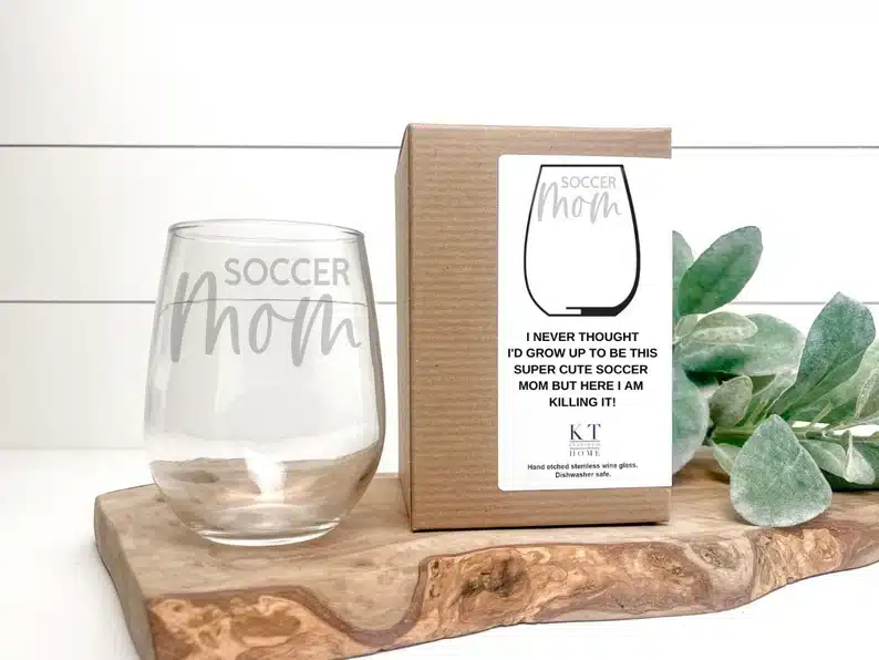 Stemless wine glass that says soccer mom with a box for it in the background. 