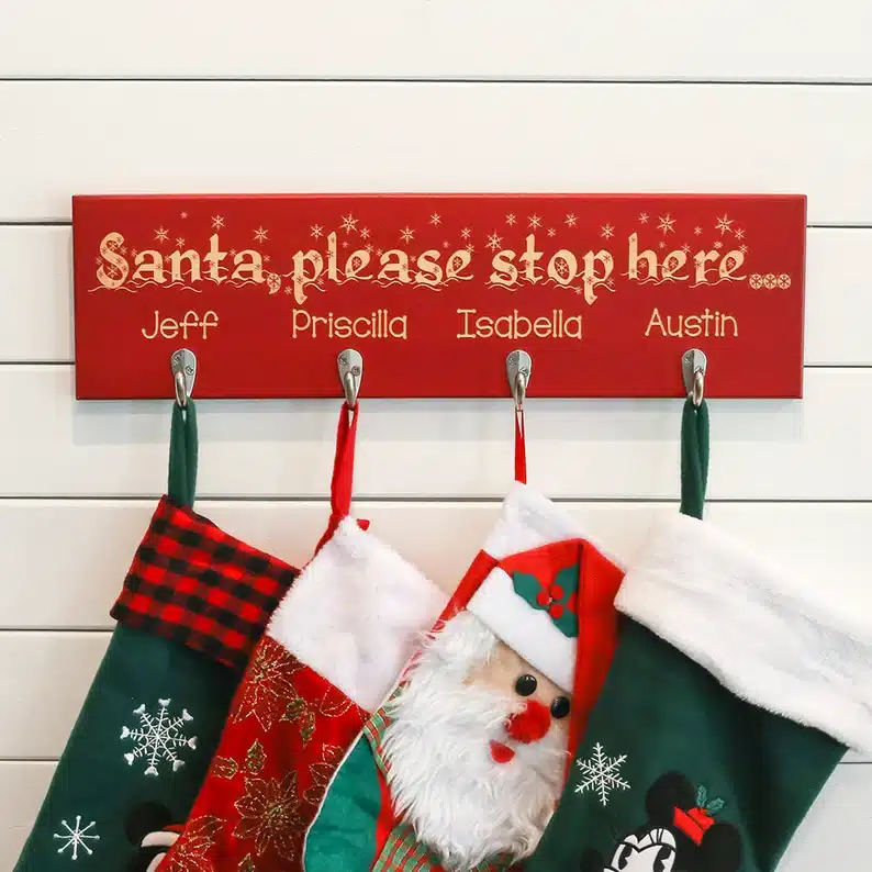 Red wooden sign personalized with family name and names of each person with a Christmas stocking hanging from it. 