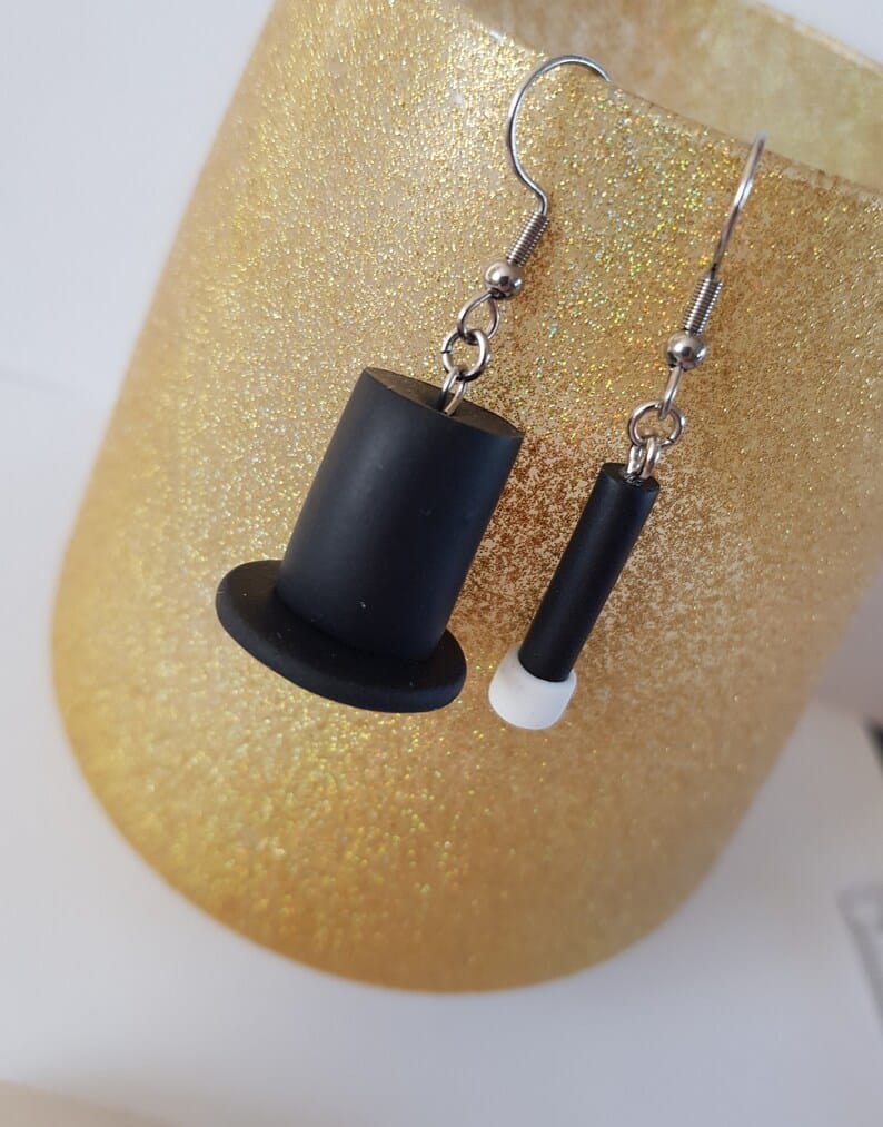 Gold background with earrings, one a black magic hat and one a black and white magic wand. 