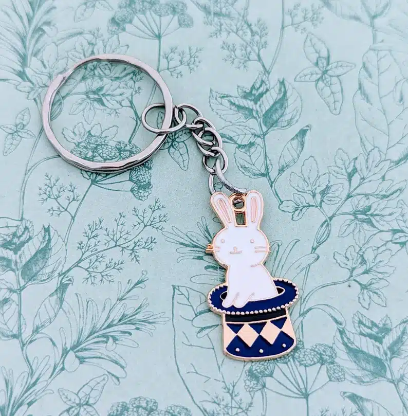 Light blue background with flowers with a silver keychain with a blue hat with a white bunny sticking out of it 