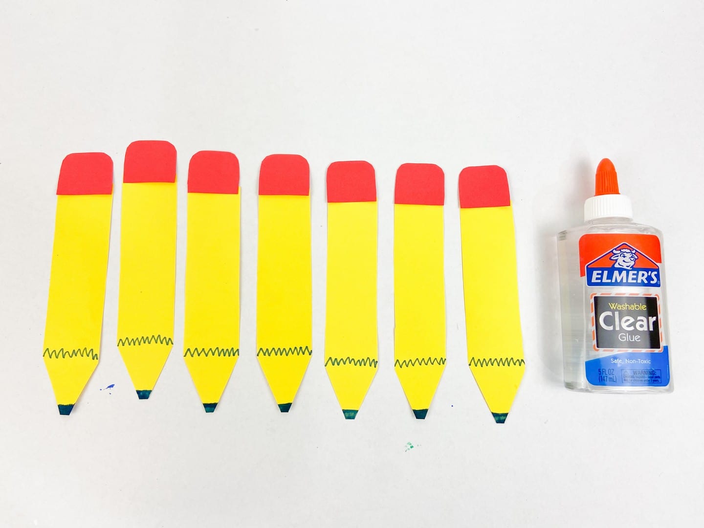 Yellow cut out pencils with red erasers cut outs glued on with a bottle of liquid glue beside it. 