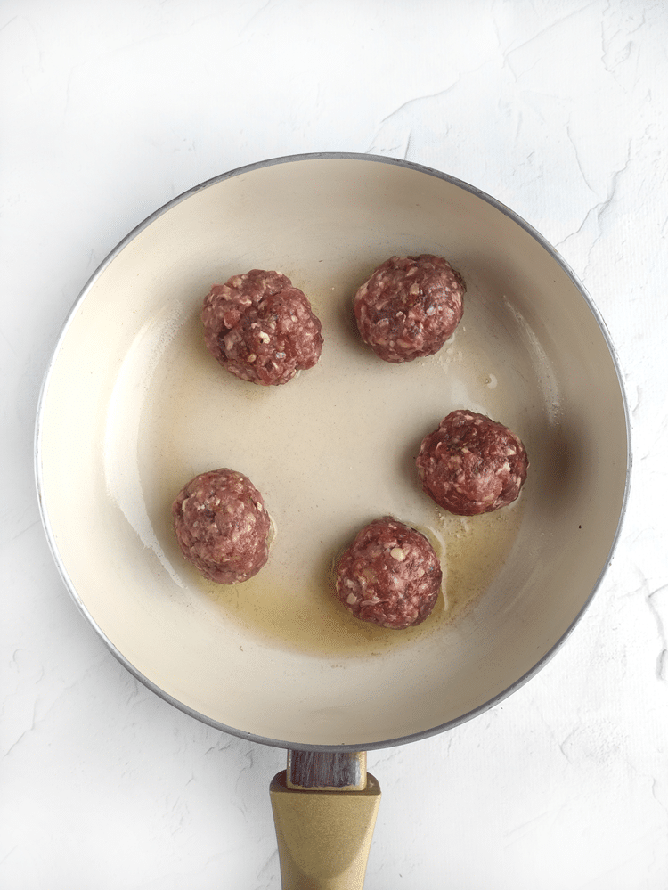 Above view of a frying pan with olive oil and five meatballs on it. 