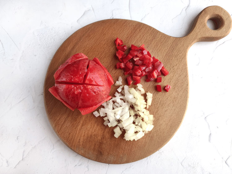 round wooden cutting board with peeled tomato, chopped up onion, and pepper. 